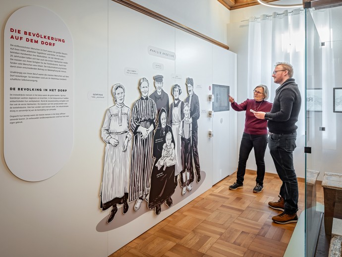 Zwei Personen schauen sich Texttafeln und die Illustration einer Familie im Apothekenmuseum Rhede an. Foto: Jannis Wiebusch © LWL